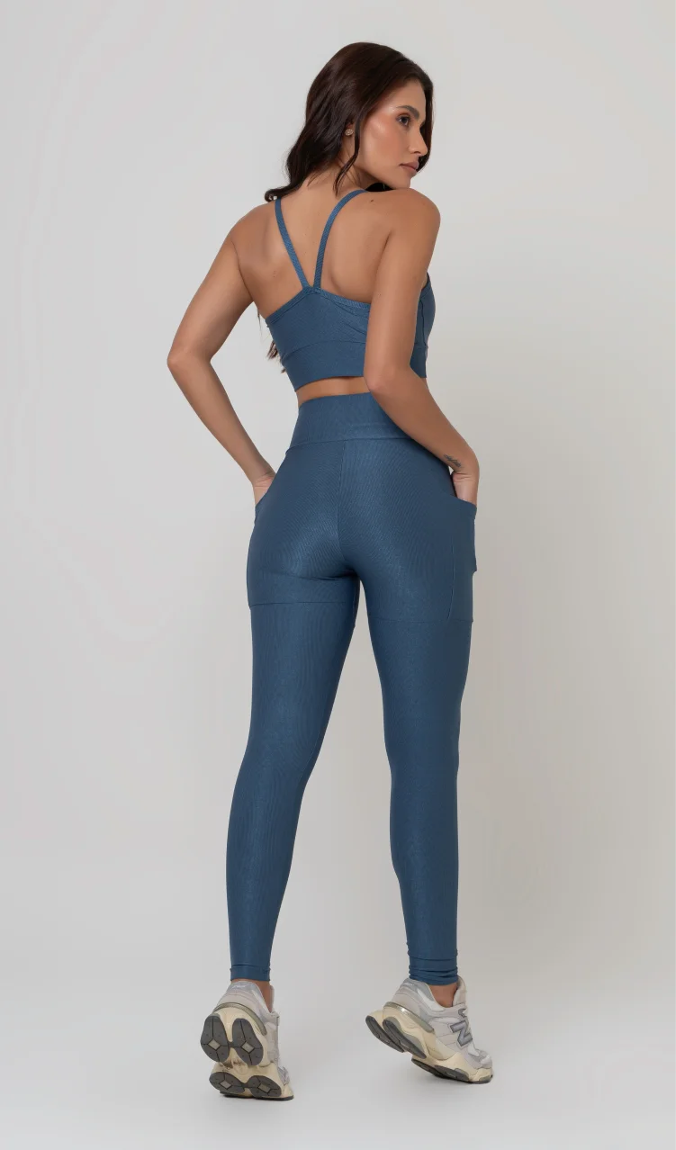 Uma foto de estúdio de corpo inteiro de uma mulher jovem posando com um conjunto fitness feminino feito de poliamida e elastano na cor safira, um tom de azul profundo, fechado e brilhante. O conjunto é composto por um top de alças finas, bojo removível e uma calça legging de cintura alta com bolsos laterais práticos. Ela está com uma mão no bolso da legging e usa tênis esportivos cinzas e bege. O fundo é cinza claro e bem iluminado. Este conjunto de treino preto é ideal para academia e exercícios físicos.