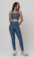 Uma foto de estúdio de corpo inteiro de uma mulher jovem posando com um conjunto fitness feminino feito de poliamida e elastano na cor safira, um tom de azul profundo, fechado e brilhante. O conjunto é composto por um top de alças finas, bojo removível e uma calça legging de cintura alta com bolsos laterais práticos. Ela está com uma mão no bolso da legging e usa tênis esportivos cinzas e bege. O fundo é cinza claro e bem iluminado. Este conjunto de treino preto é ideal para academia e exercícios físicos.