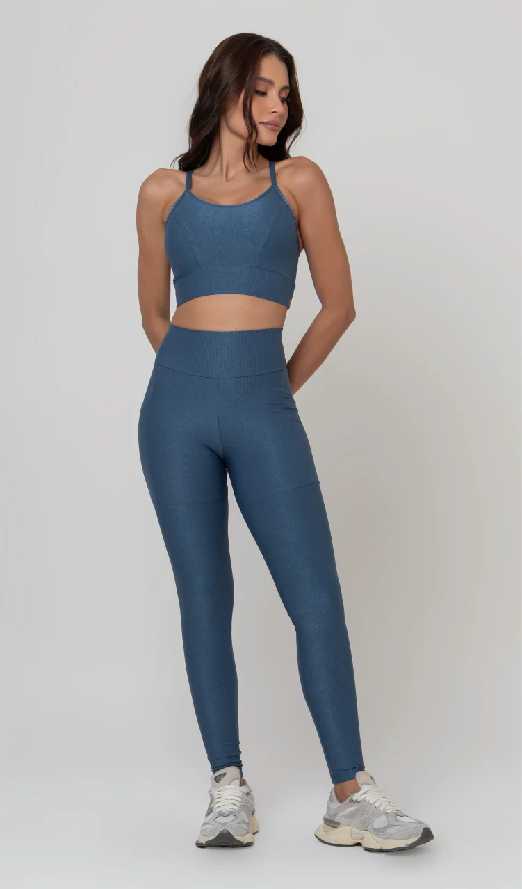 Uma foto de estúdio de corpo inteiro de uma mulher jovem posando com um conjunto fitness feminino feito de poliamida e elastano na cor safira, um tom de azul profundo, fechado e brilhante. O conjunto é composto por um top de alças finas, bojo removível e uma calça legging de cintura alta com bolsos laterais práticos. Ela está com uma mão no bolso da legging e usa tênis esportivos cinzas e bege. O fundo é cinza claro e bem iluminado. Este conjunto de treino preto é ideal para academia e exercícios físicos.