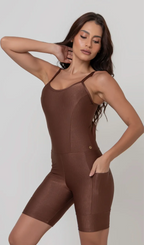 Uma foto de uma modelo feminina em pé, de frente, contra um fundo cinza claro de estúdio. Ela veste um macaquinho fitness na cor marrom, de poliamida e elastano, peça única, com alças finas, bojo e pernas curtas (estilo bermuda ciclista ou biker short). O macaquinho possui bolsos laterais práticos, proteção solar e a modelo está posando com a mão direita dentro de um deles.  O tecido parece ser elástico e confortável, ideal para treinos e academia. 