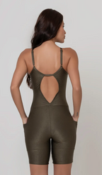 Uma foto de uma modelo feminina em pé, de costas, contra um fundo cinza claro de estúdio. Ela veste um macaquinho fitness na cor verde, de poliamida e elastano, peça única, com alças finas, bojo e pernas curtas (estilo bermuda ciclista ou biker short). O macaquinho possui bolsos laterais práticos, proteção solar e a modelo está posando com a mão direita dentro de um deles.  O tecido parece ser elástico e confortável, ideal para treinos e academia. 