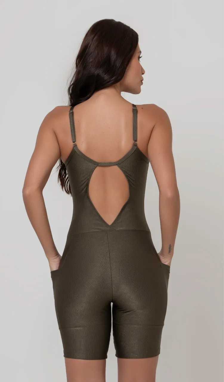 Uma foto de uma modelo feminina em pé, de costas, contra um fundo cinza claro de estúdio. Ela veste um macaquinho fitness na cor verde, de poliamida e elastano, peça única, com alças finas, bojo e pernas curtas (estilo bermuda ciclista ou biker short). O macaquinho possui bolsos laterais práticos, proteção solar e a modelo está posando com a mão direita dentro de um deles.  O tecido parece ser elástico e confortável, ideal para treinos e academia. 