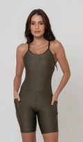 Uma foto de uma modelo feminina em pé, de frente, contra um fundo cinza claro de estúdio. Ela veste um macaquinho fitness na cor verde, de poliamida e elastano, peça única, com alças finas, bojo e pernas curtas (estilo bermuda ciclista ou biker short). O macaquinho possui bolsos laterais práticos, proteção solar e a modelo está posando com a mão direita dentro de um deles.  O tecido parece ser elástico e confortável, ideal para treinos e academia. 