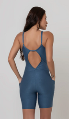 Uma foto de uma modelo feminina em pé, de costas, contra um fundo cinza claro de estúdio. Ela veste um macaquinho fitness cinza, de poliamida e elastano, peça única, com alças finas, bojo e pernas curtas (estilo bermuda ciclista ou biker short). O macaquinho possui bolsos laterais práticos, proteção solar e a modelo está posando com a mão direita dentro de um deles.  O tecido parece ser elástico e confortável, ideal para treinos e academia. 