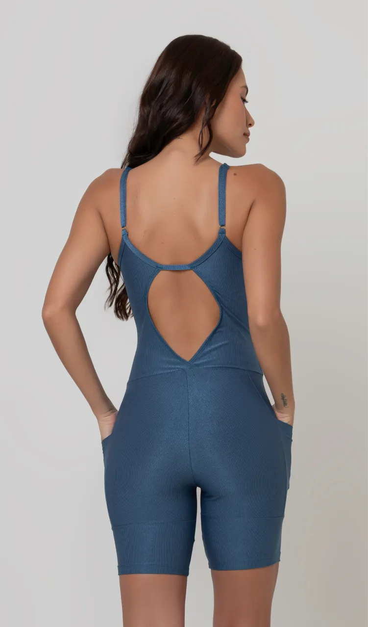 Uma foto de uma modelo feminina em pé, de costas, contra um fundo cinza claro de estúdio. Ela veste um macaquinho fitness cinza, de poliamida e elastano, peça única, com alças finas, bojo e pernas curtas (estilo bermuda ciclista ou biker short). O macaquinho possui bolsos laterais práticos, proteção solar e a modelo está posando com a mão direita dentro de um deles.  O tecido parece ser elástico e confortável, ideal para treinos e academia. 