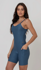 Uma foto de uma modelo feminina em pé, de frente, contra um fundo cinza claro de estúdio. Ela veste um macaquinho fitness cinza, de poliamida e elastano, peça única, com alças finas, bojo e pernas curtas (estilo bermuda ciclista ou biker short). O macaquinho possui bolsos laterais práticos, proteção solar e a modelo está posando com a mão direita dentro de um deles.  O tecido parece ser elástico e confortável, ideal para treinos e academia. 