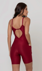 Uma foto de corpo inteiro de uma modelo feminina em pé, de costas, contra um fundo cinza claro de estúdio. Ela veste um macaquinho fitness vermelho de poliamida e elastano, peça única, com alças finas, bojo e pernas curtas (estilo bermuda ciclista ou biker short). O macaquinho possui bolsos laterais práticos, proteção solar e a modelo está posando com a mão direita dentro de um deles.  O tecido parece ser elástico e confortável, ideal para treinos e academia. 