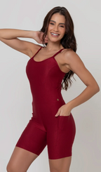 Uma foto de corpo inteiro de uma modelo feminina em pé, de frente, contra um fundo cinza claro de estúdio. Ela veste um macaquinho fitness vermelho de poliamida e elastano, peça única, com alças finas, bojo e pernas curtas (estilo bermuda ciclista ou biker short). O macaquinho possui bolsos laterais práticos, proteção solar e a modelo está posando com a mão direita dentro de um deles.  O tecido parece ser elástico e confortável, ideal para treinos e academia. 
