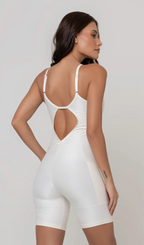 Uma foto de uma modelo feminina em pé, de costas, contra um fundo cinza claro de estúdio. Ela veste um macaquinho fitness branco, de poliamida e elastano, peça única, com alças finas, bojo e pernas curtas (estilo bermuda ciclista ou biker short). O macaquinho possui bolsos laterais práticos, proteção solar e a modelo está posando com a mão direita dentro de um deles.  O tecido parece ser elástico e confortável, ideal para treinos e academia. 