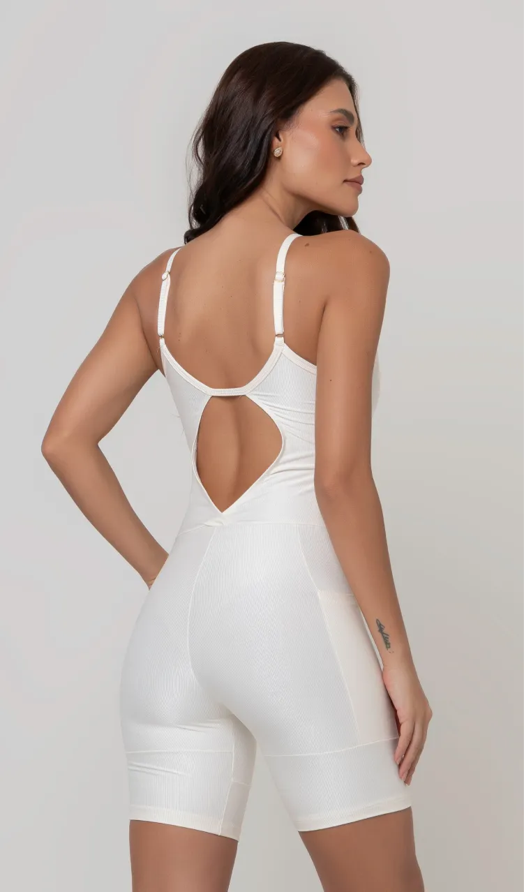 Uma foto de uma modelo feminina em pé, de costas, contra um fundo cinza claro de estúdio. Ela veste um macaquinho fitness branco, de poliamida e elastano, peça única, com alças finas, bojo e pernas curtas (estilo bermuda ciclista ou biker short). O macaquinho possui bolsos laterais práticos, proteção solar e a modelo está posando com a mão direita dentro de um deles.  O tecido parece ser elástico e confortável, ideal para treinos e academia. 