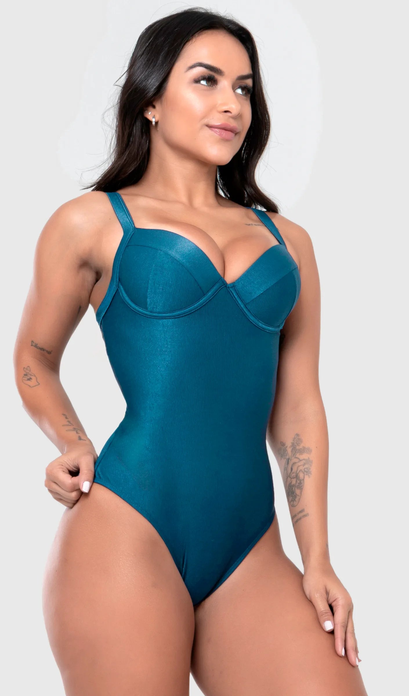 Uma modelo de cabelos escuros posa, de frente, em um estúdio com fundo cinza claro, vestindo um maiô modelador liso azul. A peça é confeccionada em tecido de poliamida e elastano, com proteção solar. Maiô estruturado com sutiã meia taça com aro para sustentação, bojo,  decote coração e alças médias. 