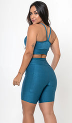 Uma modelo feminina posando de costas contra um fundo cinza neutro, vestindo um conjunto fitness feminino azul, com proteção solar. O top fitness de academia tem alças finas. A peça de baixo é um short de ciclismo (bermuda fitness feminina) de cintura alta também na cor azul, com um bolso lateral visível onde a modelo está com a mão direita inserida. Ela olha para o lado. 