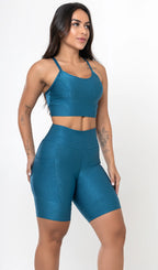 Uma modelo feminina posando de frente contra um fundo cinza neutro, vestindo um conjunto fitness feminino azul, com proteção solar. O top fitness de academia tem alças finas. A peça de baixo é um short de ciclismo (bermuda fitness feminina) de cintura alta também na cor azul, com um bolso lateral visível onde a modelo está com a mão direita inserida. Ela olha para o lado. 