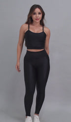 Vídeo de modelo feminina usando conjunto fitness feminino preto Mi Raromi de duas peças. Inclui um top cropped de alcinhas finas e leggings de cintura alta correspondentes, ambos feitos de poliamida e elastano. A modelo está em pé contra um fundo cinza neutro, usando tênis brancos.