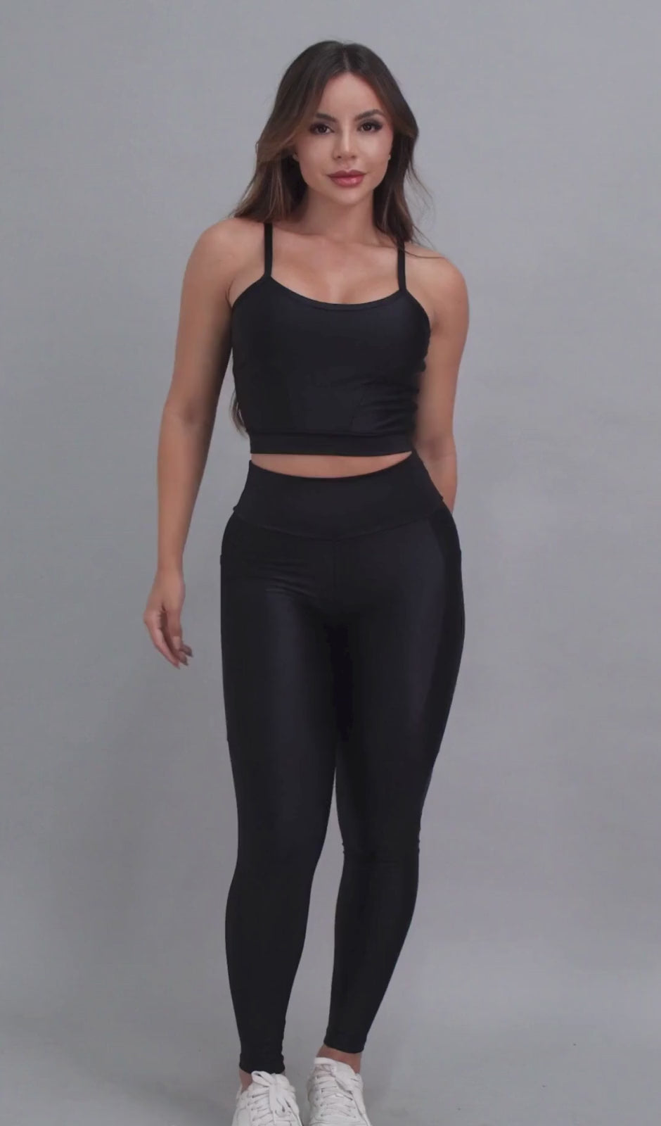 Vídeo de modelo feminina usando conjunto fitness feminino preto Mi Raromi de duas peças. Inclui um top cropped de alcinhas finas e leggings de cintura alta correspondentes, ambos feitos de poliamida e elastano. A modelo está em pé contra um fundo cinza neutro, usando tênis brancos.