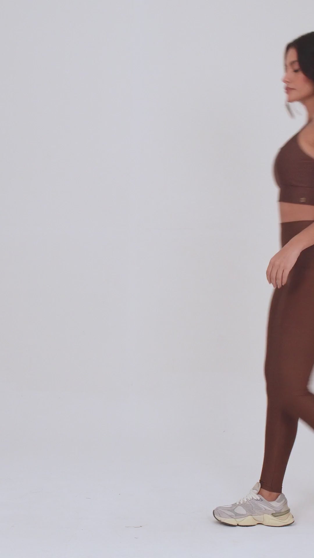Uma vídeo de estúdio de corpo inteiro de uma mulher jovem posando com um conjunto fitness feminino feito de poliamida e elastano na cor marrom. O conjunto é composto por um top de alças finas, bojo removível e uma calça legging de cintura alta com bolsos laterais práticos. Ela está com uma mão no bolso da legging e usa tênis esportivos cinzas e bege. O fundo é cinza claro e bem iluminado. Este conjunto de treino preto é ideal para academia e exercícios físicos.