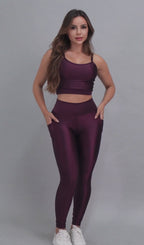 Vídeo de modelo feminina usando conjunto fitness feminino roxo uva Mi Raromi de duas peças. Inclui um top cropped de alcinhas finas e leggings de cintura alta correspondentes, ambos feitos de poliamida e elastano. A modelo está em pé contra um fundo cinza neutro, usando tênis brancos.