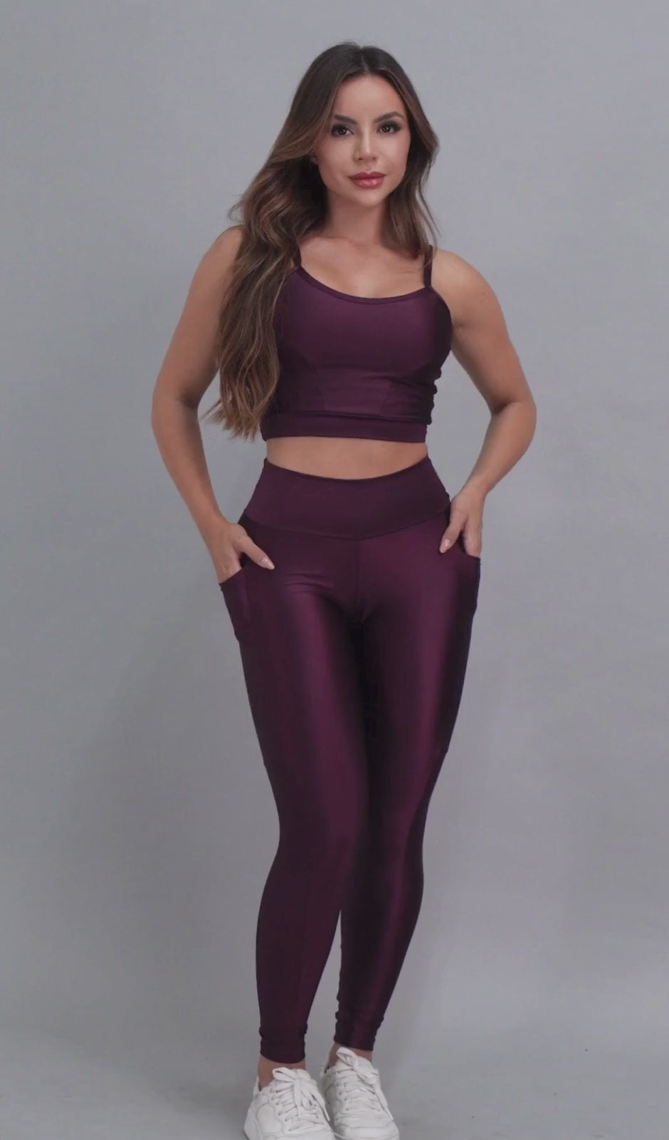 Vídeo de modelo feminina usando conjunto fitness feminino roxo uva Mi Raromi de duas peças. Inclui um top cropped de alcinhas finas e leggings de cintura alta correspondentes, ambos feitos de poliamida e elastano. A modelo está em pé contra um fundo cinza neutro, usando tênis brancos.