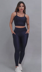 Vídeo mostrando modelo feminina usando conjunto fitness feminino azul marinho Mi Raromi de duas peças. Inclui um top cropped de alcinhas finas e leggings de cintura alta correspondentes, ambos feitos de poliamida e elastano. A modelo está em pé contra um fundo cinza neutro, usando tênis brancos.