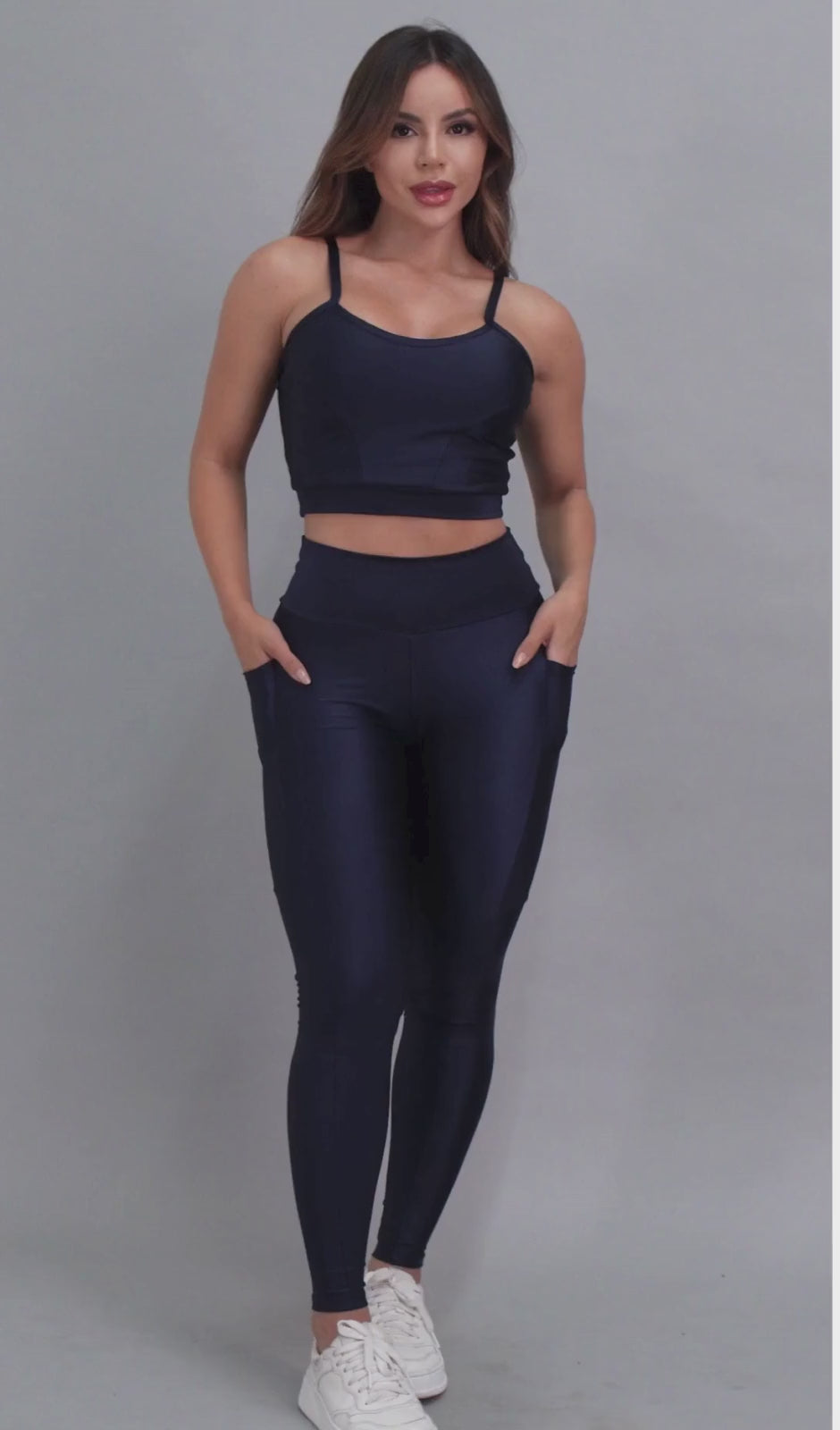 Vídeo mostrando modelo feminina usando conjunto fitness feminino azul marinho Mi Raromi de duas peças. Inclui um top cropped de alcinhas finas e leggings de cintura alta correspondentes, ambos feitos de poliamida e elastano. A modelo está em pé contra um fundo cinza neutro, usando tênis brancos.