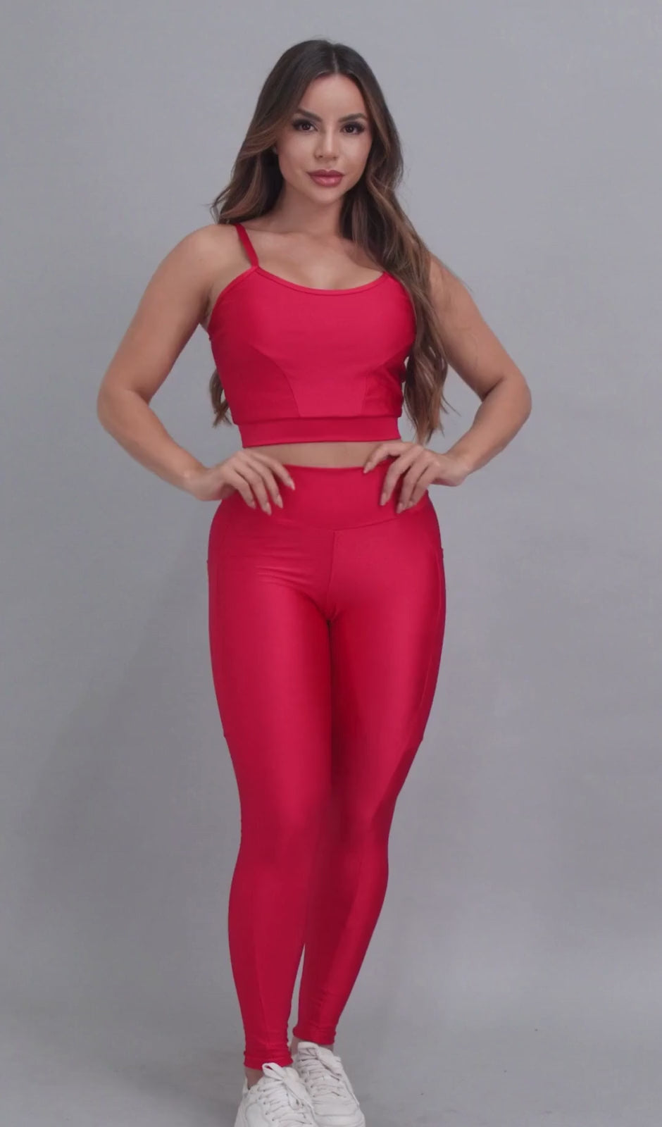 Vídeo de modelo feminina usando conjunto fitness feminino vermelho Mi Raromi de duas peças. Inclui um top cropped de alcinhas finas e leggings de cintura alta correspondentes, ambos feitos de poliamida e elastano. A modelo está em pé contra um fundo cinza neutro, usando tênis brancos.