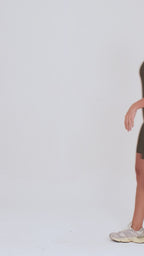 Um vídeo de uma modelo feminina em pé, de frente, contra um fundo cinza claro de estúdio. Ela veste um macaquinho fitness na cor verde, de poliamida e elastano, peça única, com alças finas, bojo e pernas curtas (estilo bermuda ciclista ou biker short). O macaquinho possui bolsos laterais práticos, proteção solar e a modelo está posando com a mão direita dentro de um deles.  O tecido parece ser elástico e confortável, ideal para treinos e academia. 