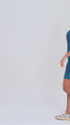 Um vídeo de uma modelo feminina em pé, contra um fundo cinza claro de estúdio. Ela veste um macaquinho fitness na cor safira, um tom de azul profundo e fechado, de poliamida e elastano, peça única, com alças finas, bojo e pernas curtas (estilo bermuda ciclista ou biker short). O macaquinho possui bolsos laterais práticos, proteção solar e a modelo está posando com a mão direita dentro de um deles.  O tecido parece ser elástico e confortável, ideal para treinos e academia. 