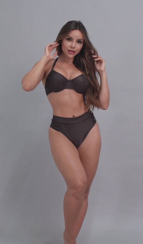 Vídeo de uma mulher modelando um conjunto de biquíni marrom escuro estruturado com aro tipo meia taça e calcinha de cintura alta texturizada,  pose de frente em estúdio contra um fundo cinza claro. 